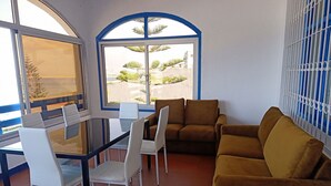 Una habitación con mesa y sillas de vidrio, un sofá marrón y un ventanal con vistas a zonas verdes y edificios.