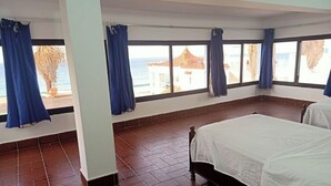 A room with a bed, large windows with curtains, a tiled floor, and a view of the sea and buildings.