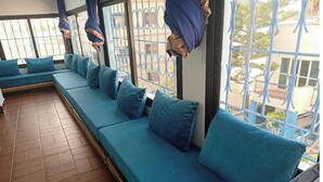 A long, blue cushioned bench with pillows, a tiled floor, and a view through a metal gate.