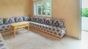 Een kamer met een beige muur, een groot raam met witte kaders, een gestreept sofa met paarse kussens, en een houten tafel.