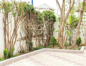 A courtyard with a tiled floor, a white wall, and various trees and shrubs.