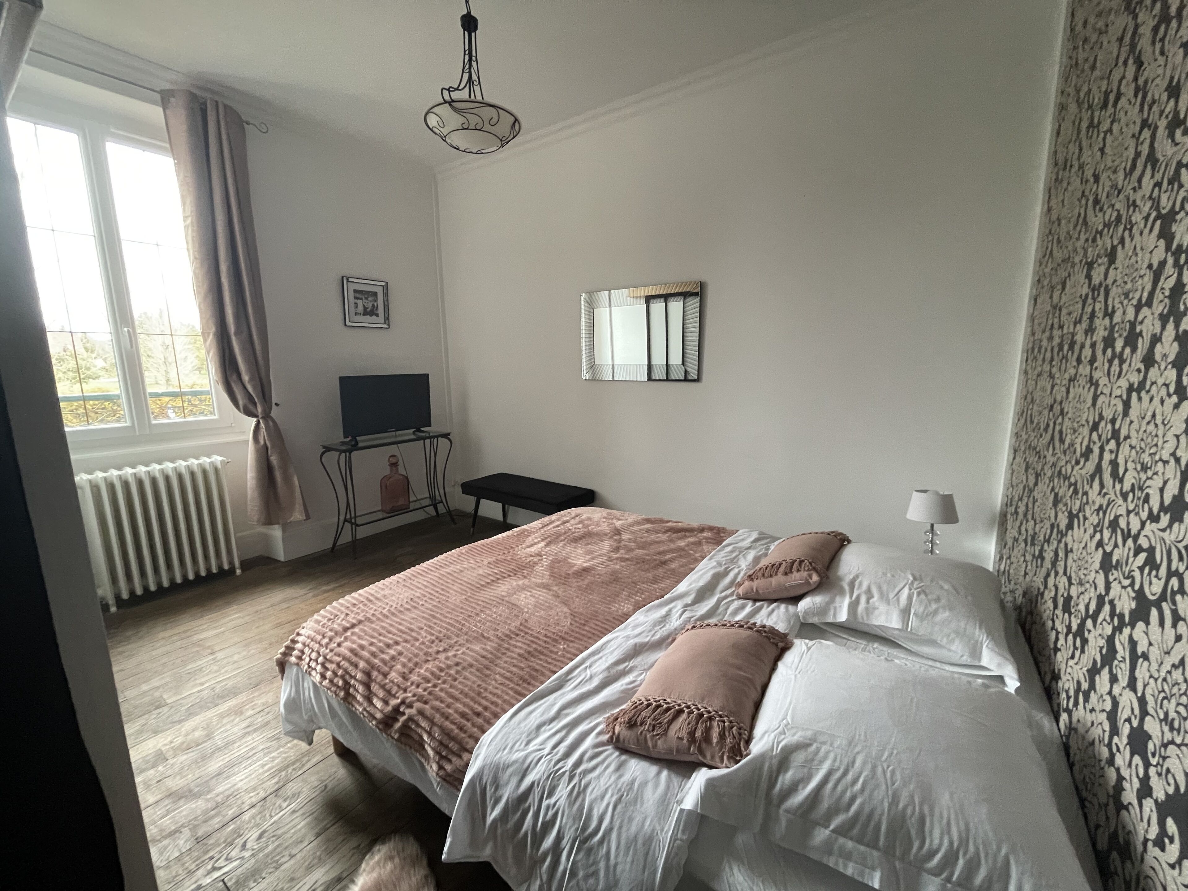 A bedroom with a bed, a small table, a television, a radiator, and a window with curtains.
