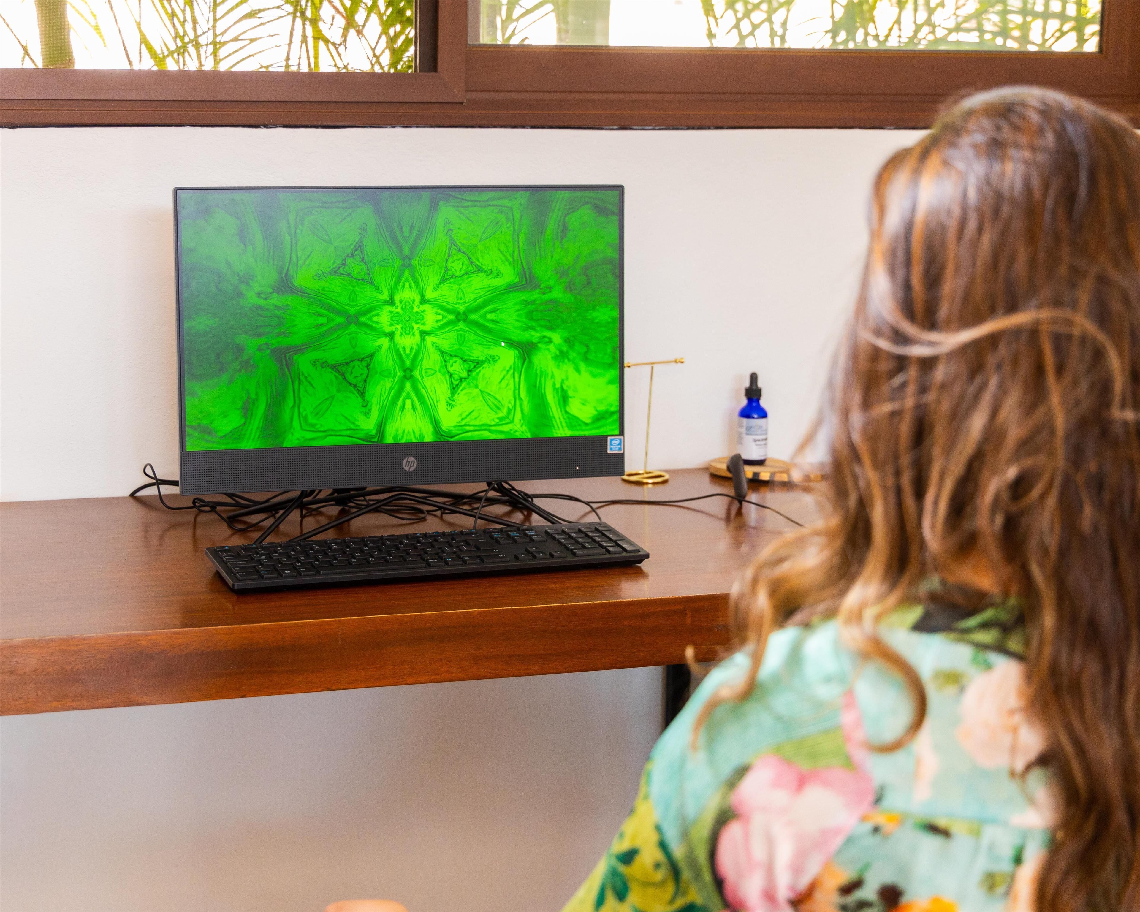 A person sitting at a wooden desk with a computer monitor displaying a green abstract image, a keyboard, and a bottle of hand sanitizer.