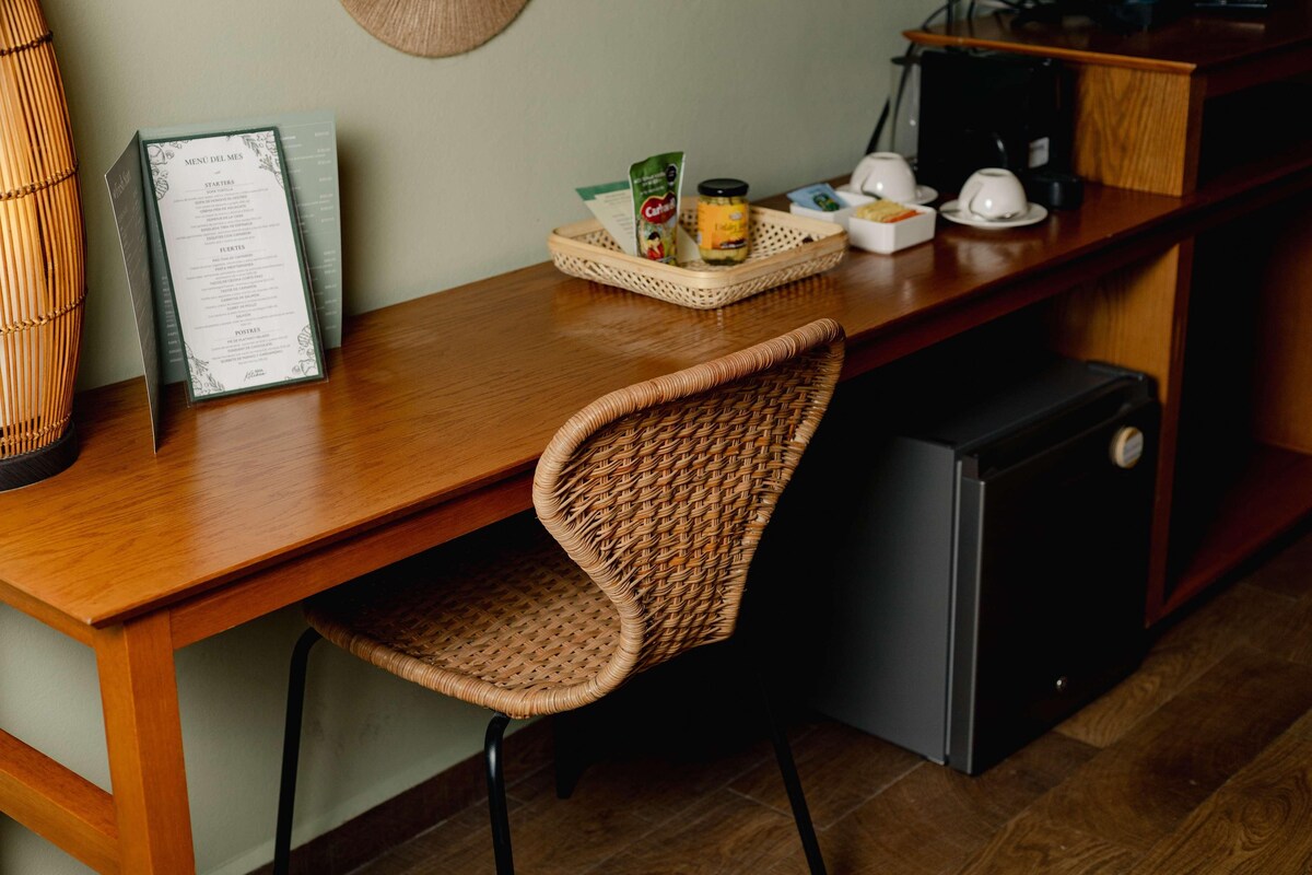 Una mesa de comedor de madera con una silla de mimbre, un menú, una cafetera y un refrigerador pequeño.