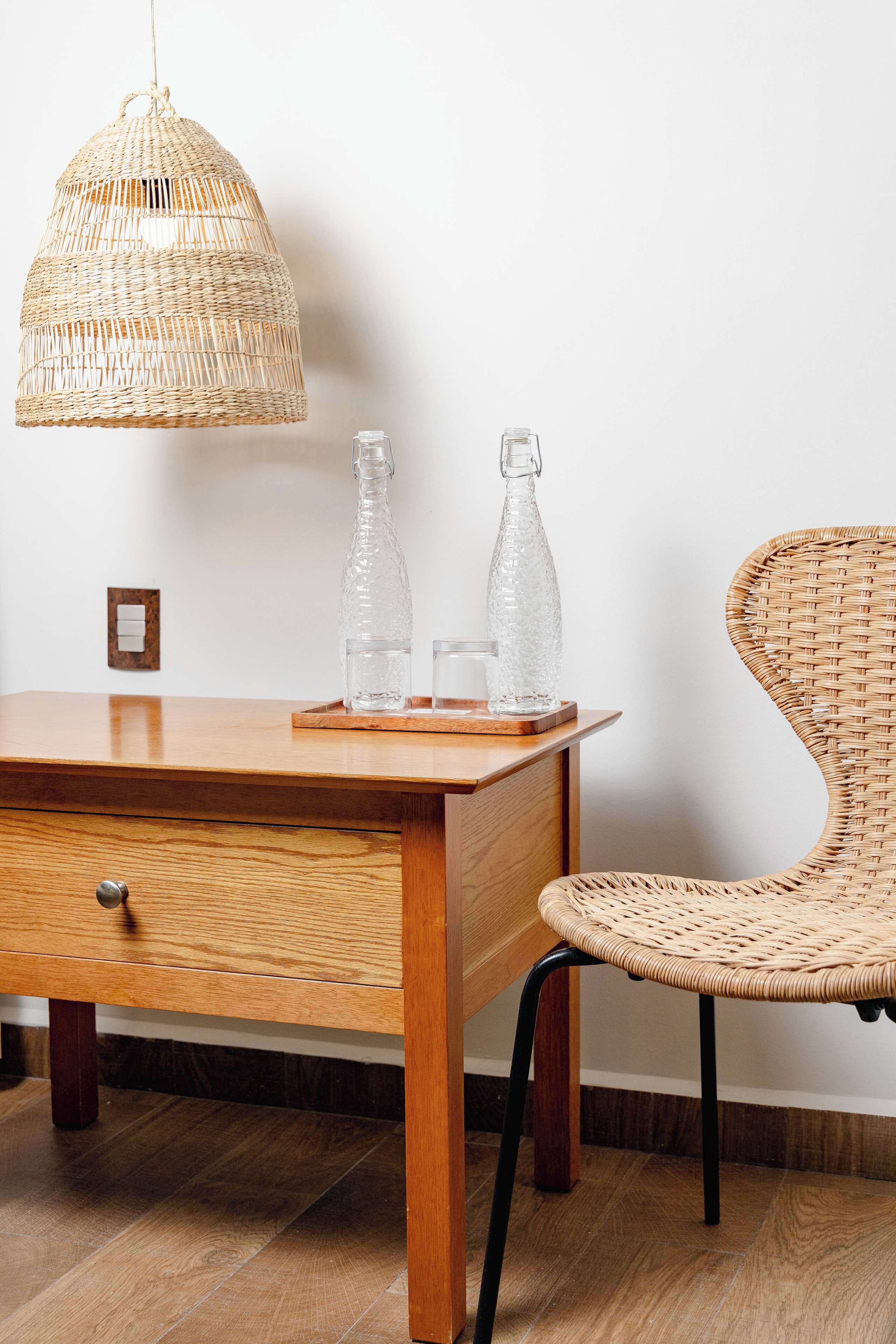 A wooden desk with a wicker chair, a hanging wicker light fixture, and two clear glass bottles on the desk.