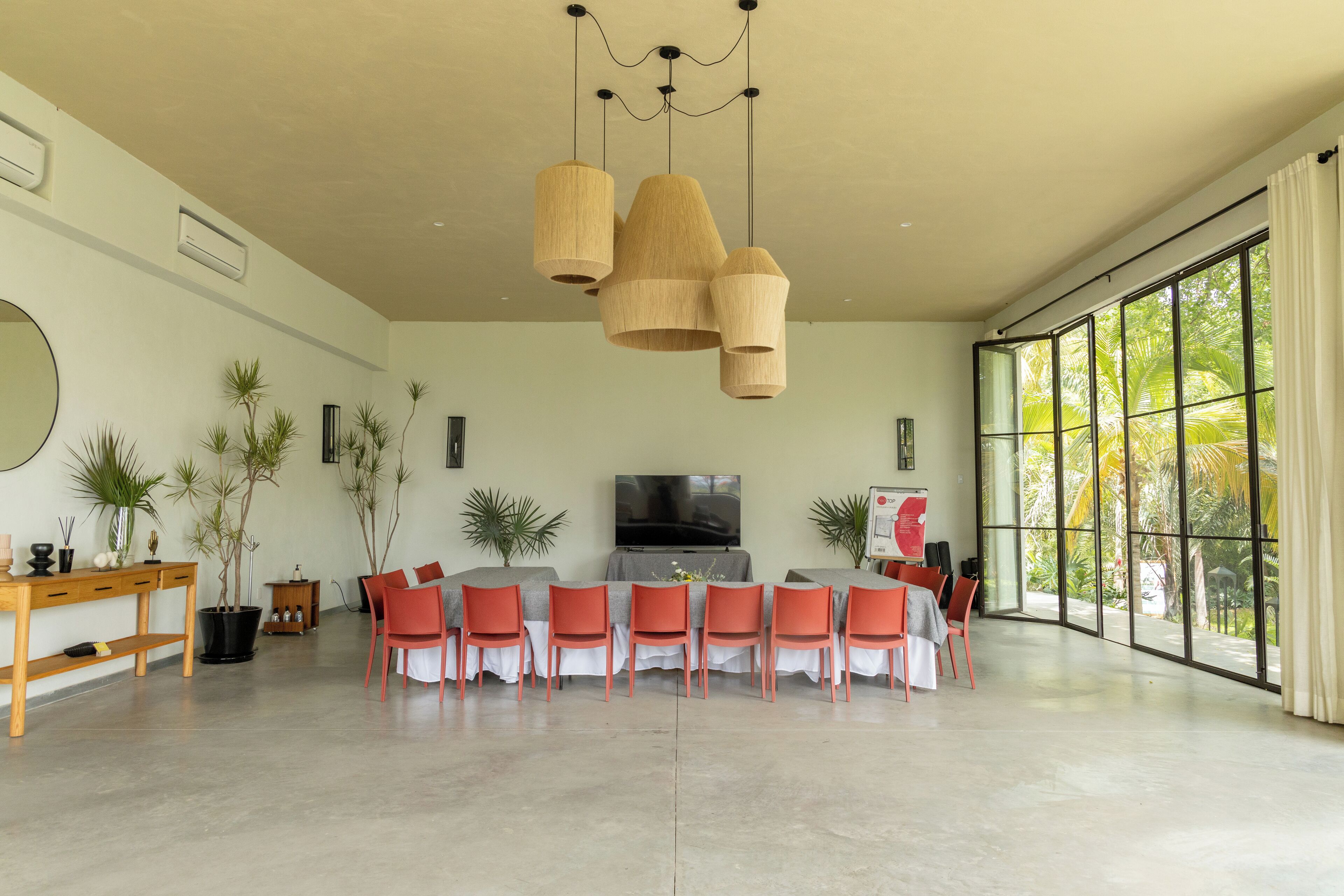 A spacious room with a long dining table, multiple chairs, a television, and large windows offering a view of palm trees.