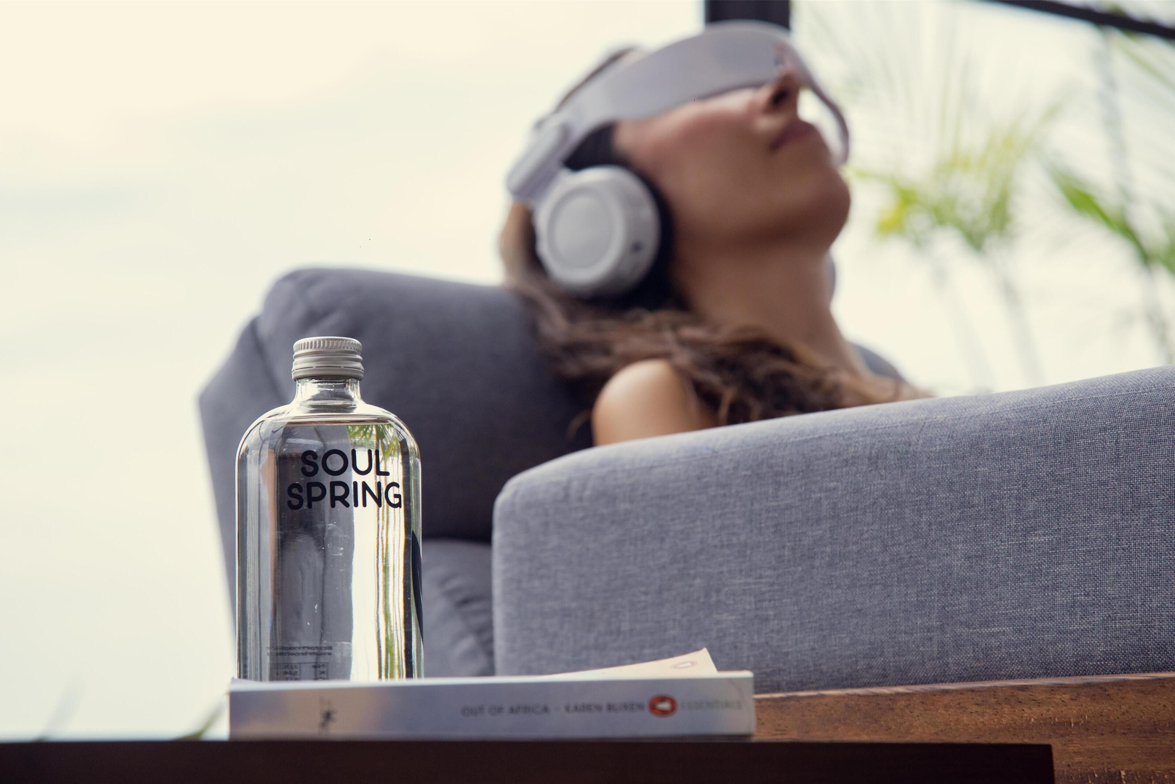 A person reclining on a grey sofa with a clear bottle labeled 'SOUL SPRING' and a book titled 'OUT OF AFRICA - LAUREN BUESEN' on a wooden table.