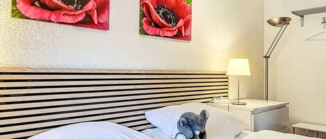 A hotel room with a striped headboard, white bedding, a lamp on the nightstand, and two red flower paintings on the wall.