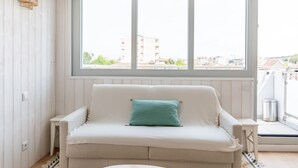 Ein Zimmer mit einem weißen Sofa, einem türkisfarbenen Kissen und einem Fenster mit Blick auf Gebäude.
