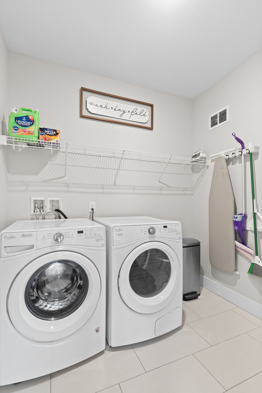A laundry room with two front-loading washing machines, a shelf with laundry detergent, a hanging rack, and a broom.