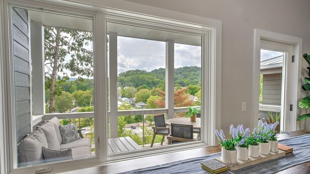 A room with a large sliding glass door leading to a balcony with a view of a green landscape and houses. There is a sofa with cushions and a small table with potted plants. - Northshore Chattanooga - Hot Tub - Walk to Dining, Publix & Coolidge Park (Chattanooga)