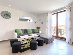 Un salon moderne avec un canapé gris, des coussins verts, une table basse blanche et des poufs noirs. On y trouve une grande fenêtre avec des rideaux, une horloge murale et des appliques murales décoratives.