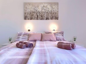 Un lit bien fait, avec du linge de lit rose et des serviettes pliées, un tableau représentant des arbres accroché au mur et des lampes de chevet.