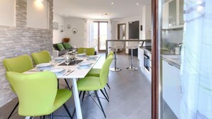 Un espace repas moderne avec une longue table, des chaises vertes et une cuisine visible à travers une porte ouverte.