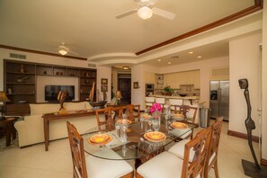 A spacious living room with a glass-top dining table set for six, surrounded by wooden chairs. A television is mounted on the wall, and there are built-in shelves with various decorative items. - Bella Vista 2D 3 bdrs pool view in Los Suenos (Playa Herradura)