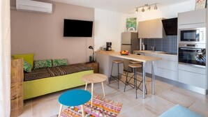 Un espacio habitable compacto con kitchenette, un sofá con cojines estampados, un televisor montado en la pared y una pequeña zona de bar con taburetes.