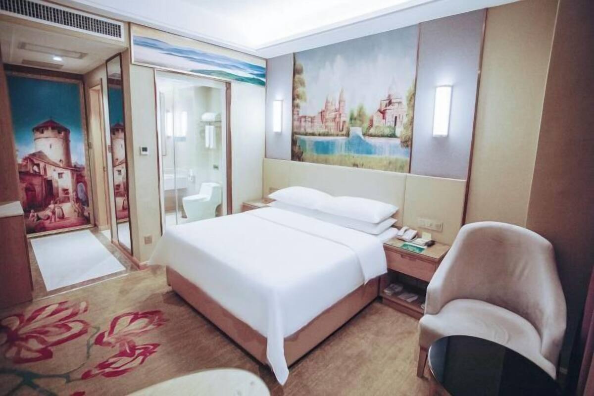 Hotellihuone, jossa on sänky, tuoli, yöpöytä, seinällä oleva taulu ja ovi, joka johtaa kylpyhuoneeseen.