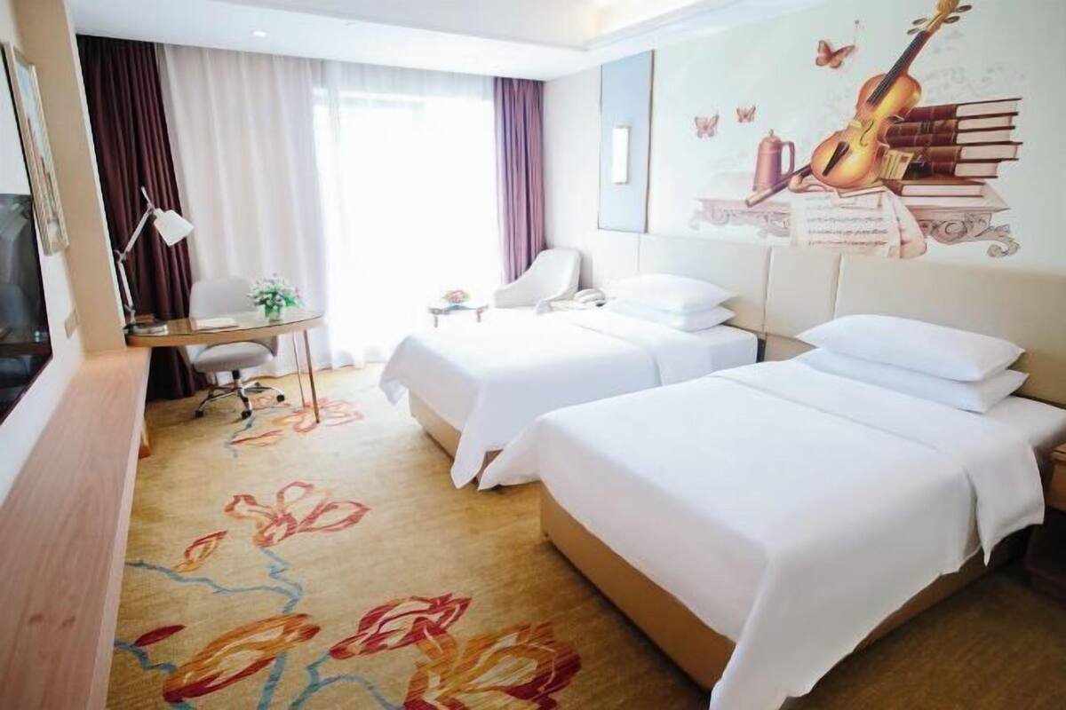 Hotellihuone, jossa on kaksi sänkyä, työpöytä, tuoli ja suuri ikkuna, jossa on verhot.