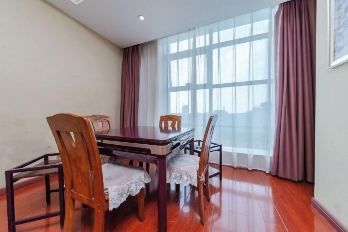 A dining area with a wooden table and chairs, a lace tablecloth, and a view through large windows with sheer curtains.