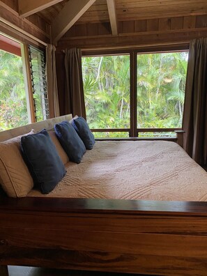 A bed with a quilted cover and four pillows, a wooden headboard, and a view of lush greenery through large windows.