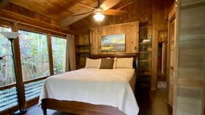 Een slaapkamer in een houten blokhut met een bed, een plafondventilator en een groot raam.