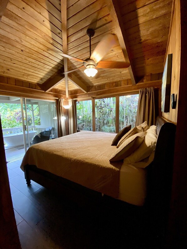A spacious bedroom with a large bed, a ceiling fan, and a view of lush greenery through large windows.