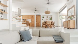Una sala de estar moderna con un sofá color beige, una mesa de comedor de madera con sillas, y una zona de cocina con armarios de madera y una lámpara colgante.