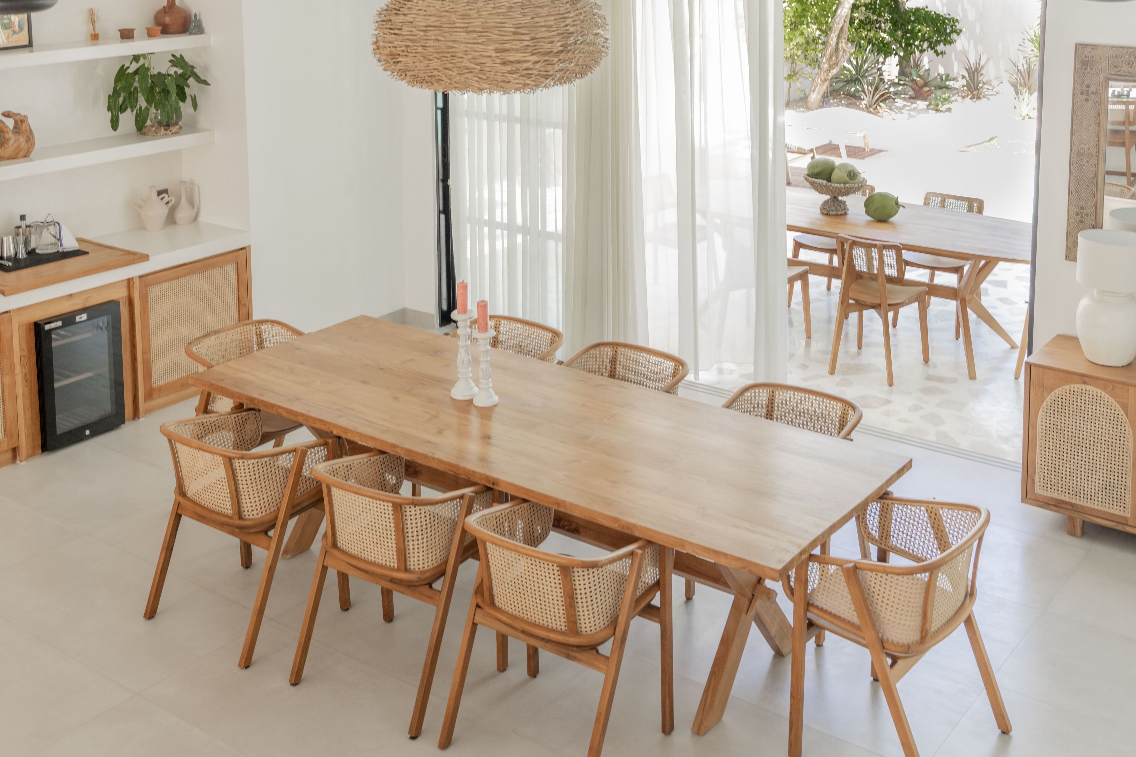 Un vaste espace dédié à la salle à manger, avec une longue table en bois et des chaises en osier, le tout entouré de murs blancs et agrémenté d’une grande fenêtre.