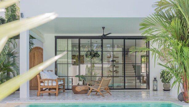 Un área de piscina moderna con dos sillones de madera, una puerta de vidrio que da a una habitación y un ventilador de techo.