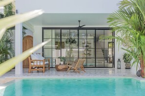 A modern pool area with two wooden lounge chairs, a glass door leading to a room, and a ceiling fan. - Casa Philip (Pecatu)