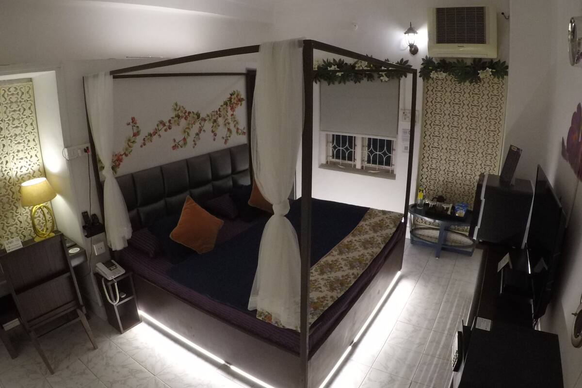 Een hemelbed met baldakijn, een klein nachtkastje met lamp, een televisie en een decoratief wandpaneel.