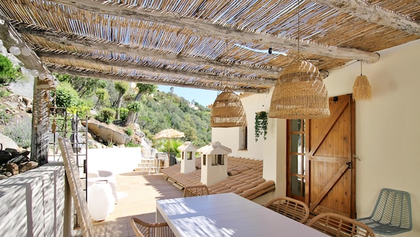Um pátio externo com uma mesa comprida e cadeiras de vime, uma porta de madeira e um telhado de palha.