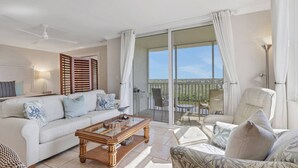 A spacious living room with a white sofa, a wooden coffee table, and a patterned armchair. A balcony with a view of the landscape is visible through large glass doors.