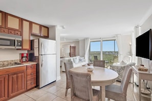 A modern kitchen with wooden cabinets, a white refrigerator, and a coffee maker. There is a dining area with wicker chairs and a round table. A large window offers a view of the outdoors.