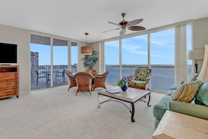 A spacious living room with a large window overlooking a body of water, a ceiling fan, and a television on a wooden cabinet.