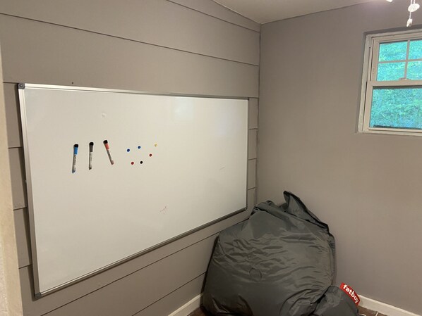 Una pizarra con marcadores y un sillón tipo bean bag gris en una habitación con paredes grises y una ventana.