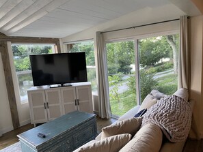 Una sala de estar con televisor sobre un mueble, un sofá y vistas al exterior con vegetación.