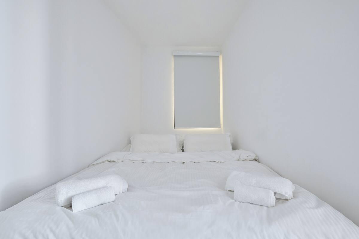 Une chambre d’hôtel au décor minimaliste, avec un grand lit, du linge de lit blanc et une table de chevet dotée d’une lampe.