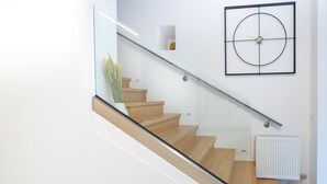 A modern staircase with glass railings and wooden steps, leading to a white wall with a minimalist clock and a radiator.