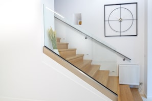 Een moderne trap met glazen balustrades en houten treden, die leidt naar een witte muur met een minimalistische klok en een radiator.