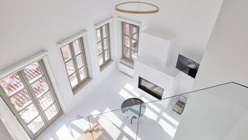 Un espacio habitable diáfano con techos altos, grandes ventanales, una escalera de cristal y una lámpara moderna.