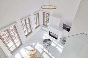 Un espacio habitable diáfano con techos altos, grandes ventanales, una escalera de cristal y una lámpara moderna.
