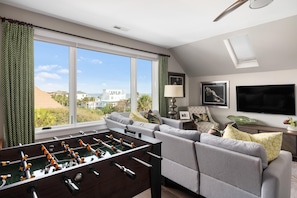A living room with a foosball table, a grey sofa, and a large window offering a view of the city.