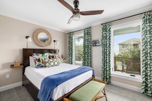 A bedroom with a bed, a ceiling fan, a window with curtains, a wooden headboard, a bedside table with a lamp, and a small shelf.