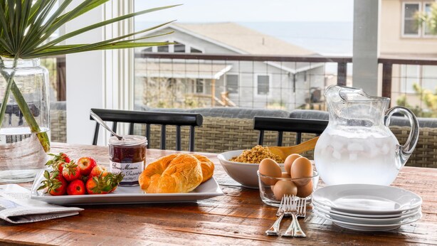 Une table de petit-déjeuner avec un croissant, des fraises, des œufs et une carafe de lait, le tout placé sur un balcon donnant sur une maison.