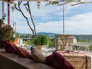 Une terrasse avec un mur en pierre, une table en bois recouverte d’une nappe rouge, et une vue sur la mer et les montagnes.