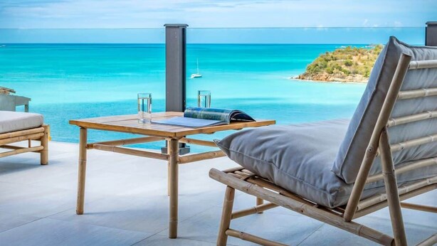 A wooden lounge chair with a cushion, a small wooden table with a glass and a book, and a view of the ocean and a small island. - Sunset Vista Suite – Panoramic Ocean Retreat at Tamarind Hills, Antigua (Ffryes beach)