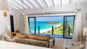 A modern living room with a large sliding glass door leading to a balcony with a view of a beach and ocean. - Sunset Vista Suite – Panoramic Ocean Retreat at Tamarind Hills, Antigua (Ffryes beach)