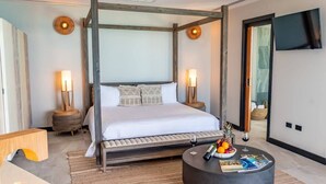 A modern bedroom with a four-poster bed, bedside lamps, a round coffee table with fruits and a wine glass, and a TV mounted on the wall. - Panorama Suite – Oceanview Luxury Suite at Tamarind Hills, Antigua (Unknown)