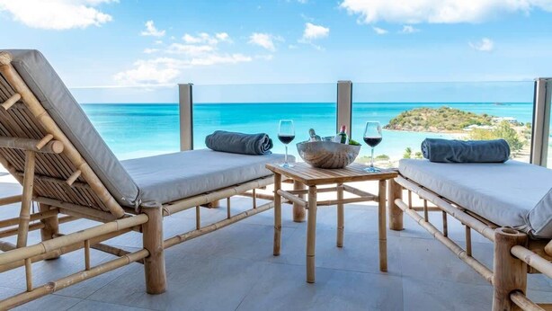 A beachside lounge with two cushioned chairs, a table with a drink and a bowl, and a view of the ocean and a small island. - Panorama Suite – Oceanview Luxury Suite at Tamarind Hills, Antigua (Unknown)